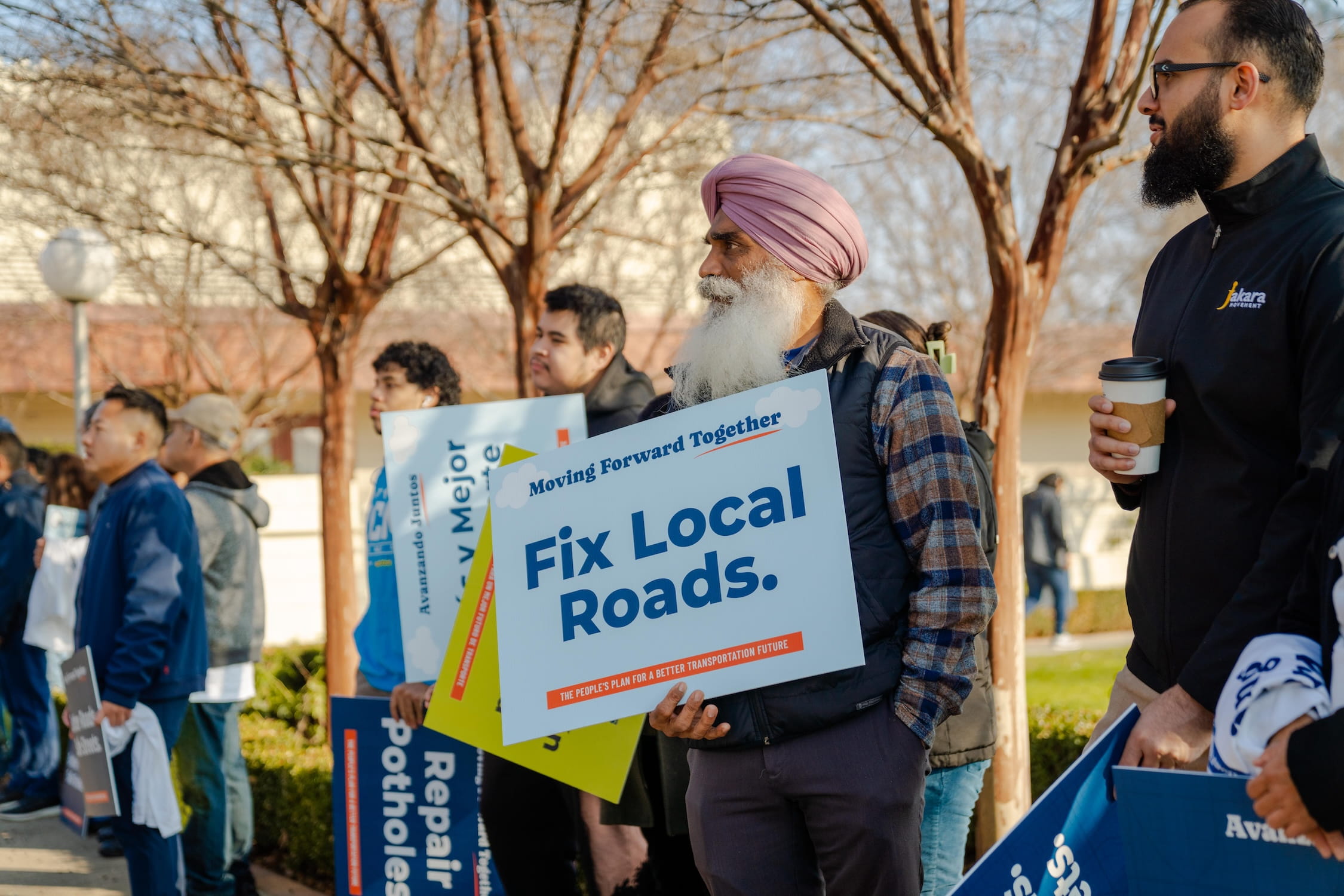 A row of supporters listening to the speaker. They are holding colorful signs, with one stating, "Fix Local Roads."