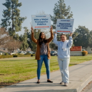 Two women holding blue signs high in the air. One sign reading, "Mas y Mejor Transporte Publico" and the other reading, "Reparemos Las Carretaras Locales."