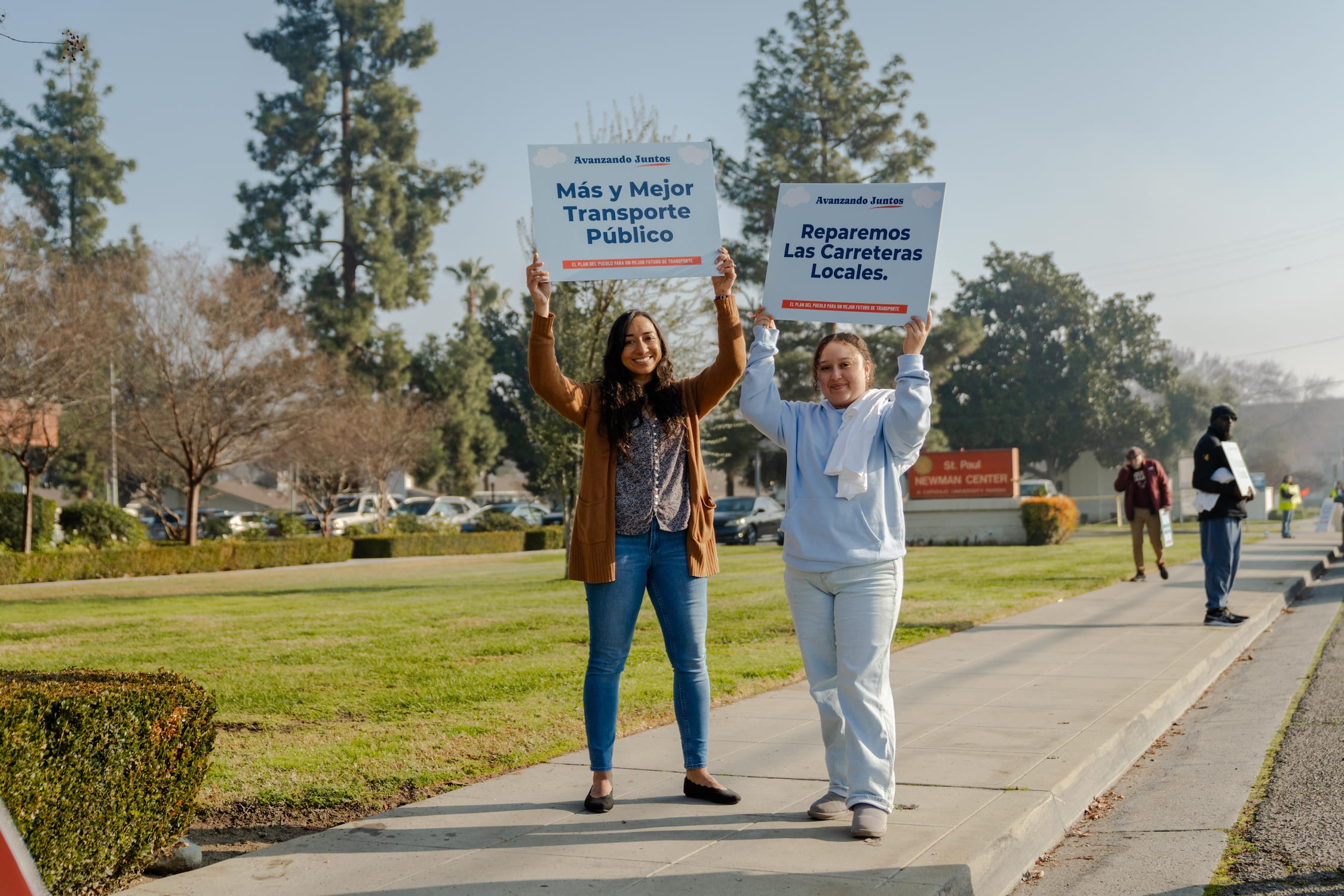 Two women holding blue signs high in the air. One sign reading, "Mas y Mejor Transporte Publico" and the other reading, "Reparemos Las Carretaras Locales."