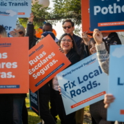 A group of supporters with their colorful signs, cheering and smiling. One person is focused on with a sign reading, "Rutas Escolares Seguras."