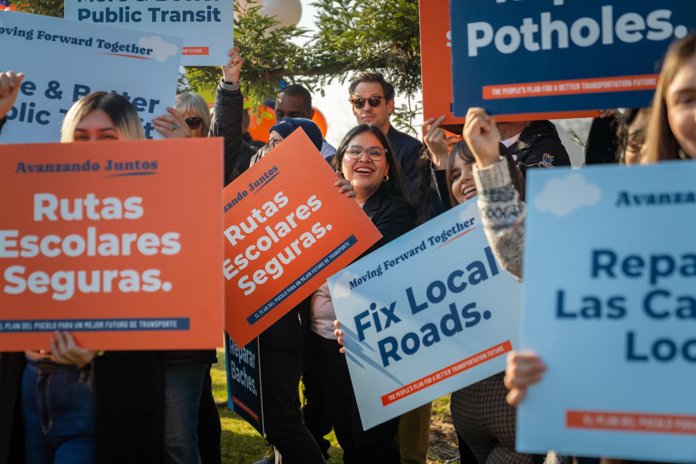 A group of supporters with their colorful signs, cheering and smiling. One person is focused on with a sign reading, "Rutas Escolares Seguras."