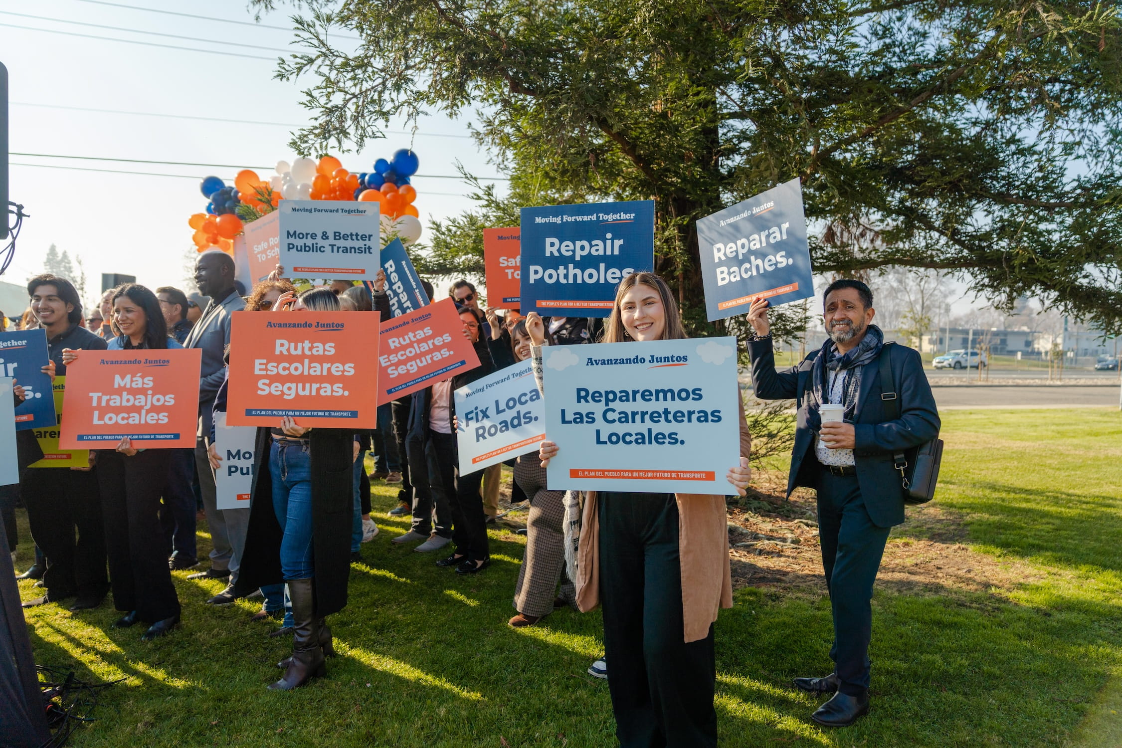 A wide-angle shot of group of people holding their colorful signs. One woman is standing directly in view holding a sign that reads, "Reparemos Las Carreteras Locales."