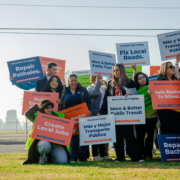 A wide-angle photo of a group of people holding their signs, listening to the speaker. Some signs reading statements like, "Repair Potholes," or "Create Local Jobs."