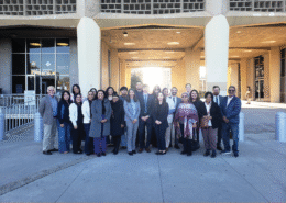 A wide group portrait of about twenty people of diverse ages and backgrounds standing in front of a modern "Court House Entrance." They are dressed in a mix of professional and business-casual attire. The courthouse features a unique concrete honeycomb-style facade.