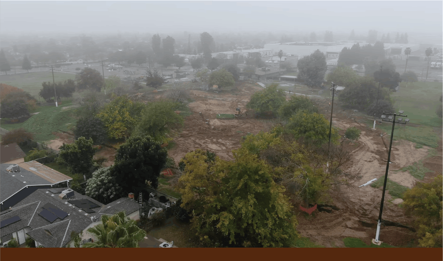 Aerial view of Calwa Park during renovation, showing a foggy construction site with earthwork underway and trees and nearby homes surrounding the area.