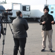 Community advocate speaks to media during a press conference supporting commercial drivers affected by California DMV license revocations, with trucks parked in the background.