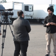 Community advocate speaks to media during a press conference supporting commercial drivers affected by California DMV license revocations, with trucks parked in the background.