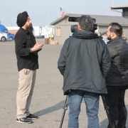 Community advocate speaks to media during a press conference supporting commercial drivers affected by California DMV license revocations, with trucks parked in the background.
