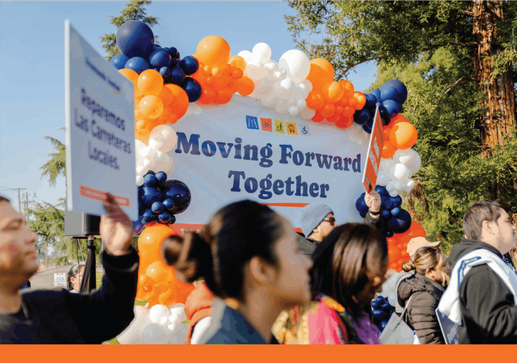 A group of approximately a dozen diverse community members and speakers stand together in front of a large orange and blue balloon arch behind them.
