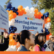 A group of approximately a dozen diverse community members and speakers stand together in front of a large orange and blue balloon arch behind them.