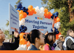A group of approximately a dozen diverse community members and speakers stand together in front of a large orange and blue balloon arch behind them.