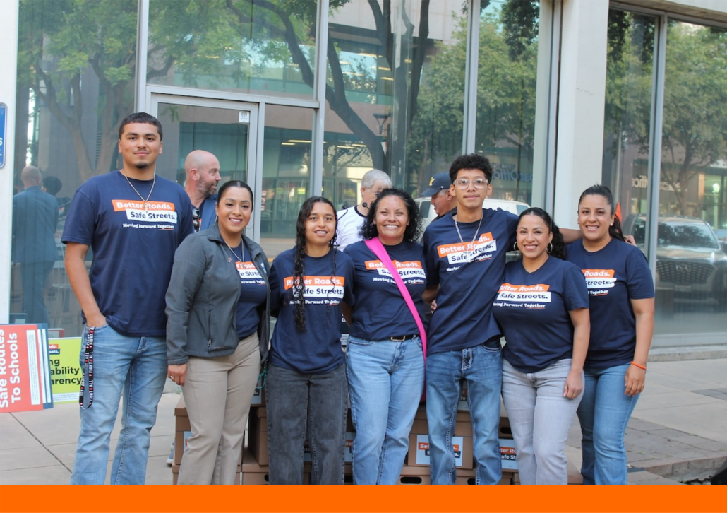 group of six smiling individuals standing outdoors in front of a glass-fronted building. They are wearing matching navy blue t-shirts that read "Better Roads. Safe Streets. Moving Forward Together." They are standing in front of a stack of several cardboard Banker boxes.