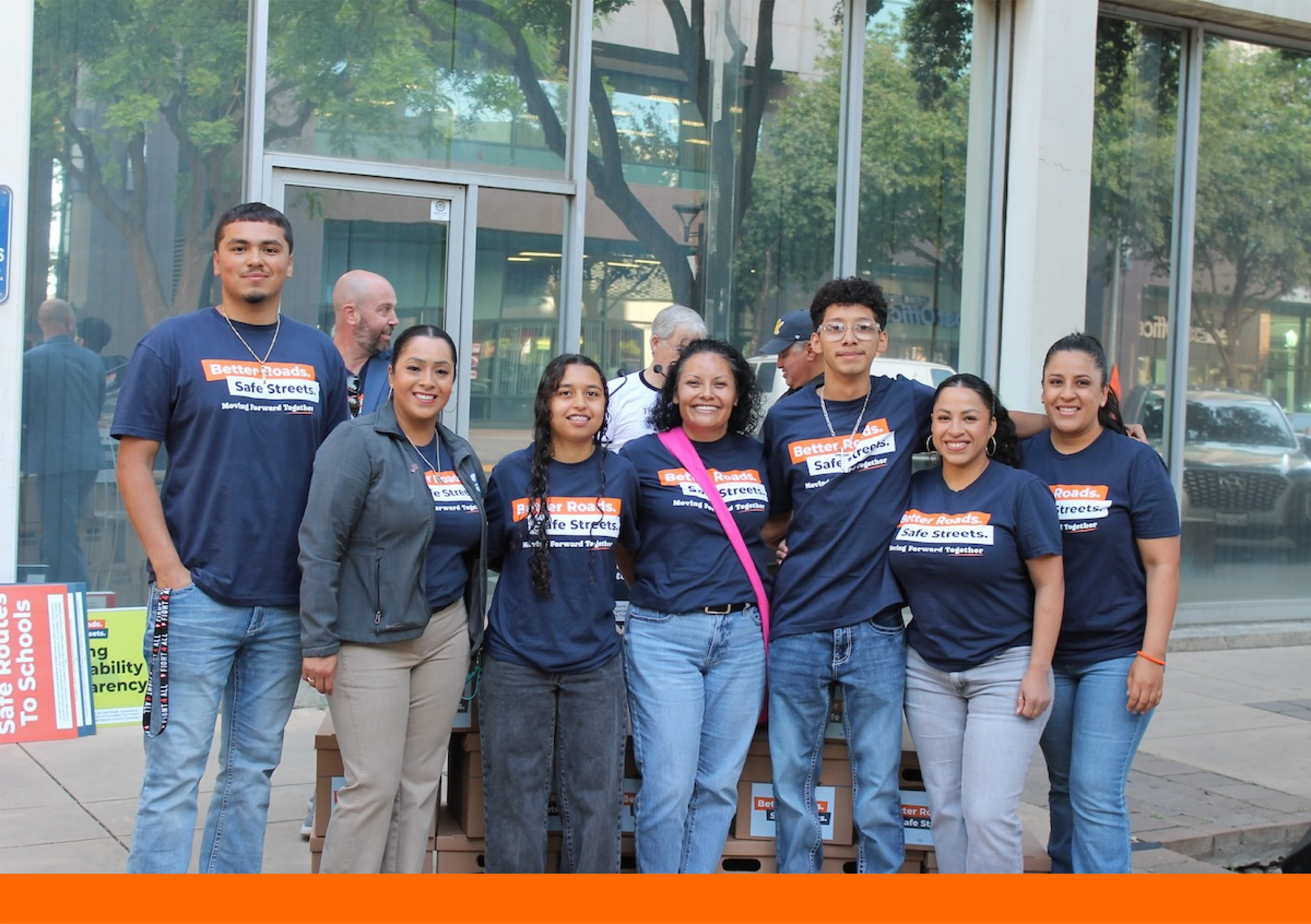 group of six smiling individuals standing outdoors in front of a glass-fronted building. They are wearing matching navy blue t-shirts that read "Better Roads. Safe Streets. Moving Forward Together." They are standing in front of a stack of several cardboard Banker boxes.