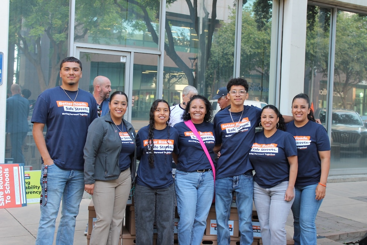 A group of six smiling individuals standing outdoors in front of a glass-fronted building. They are wearing matching navy blue t-shirts that read 