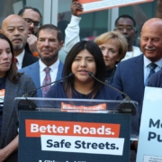 A young woman with braces speaking into microphones at a clear podium. The podium has a sign reading "Better Roads. Safe Streets. A Citizen-Led Effort." She is surrounded by a large, supportive crowd of diverse individuals holding campaign signs, some wearing campaign t-shirts and others in business attire.