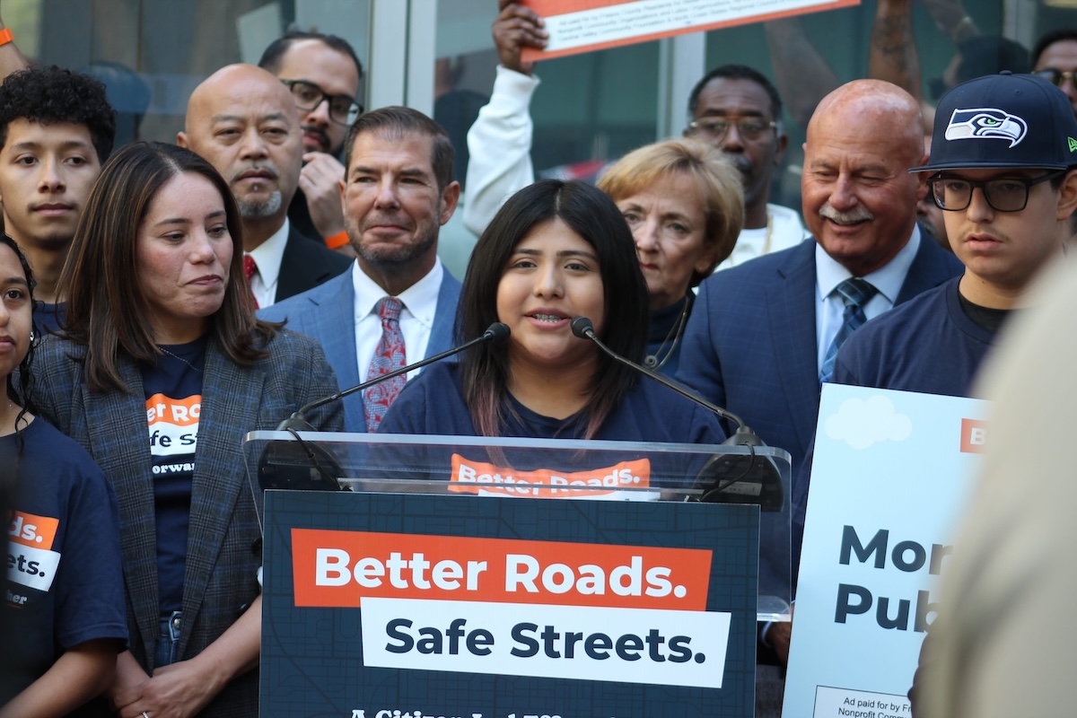 A young woman with braces speaking into microphones at a clear podium. The podium has a sign reading "Better Roads. Safe Streets. A Citizen-Led Effort." She is surrounded by a large, supportive crowd of diverse individuals holding campaign signs, some wearing campaign t-shirts and others in business attire.