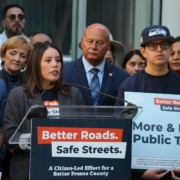A woman wearing a plaid blazer over a navy campaign t-shirt speaking at the "Better Roads. Safe Streets." podium. She is surrounded by the same crowd of supporters and campaign members, who are watching her speak.