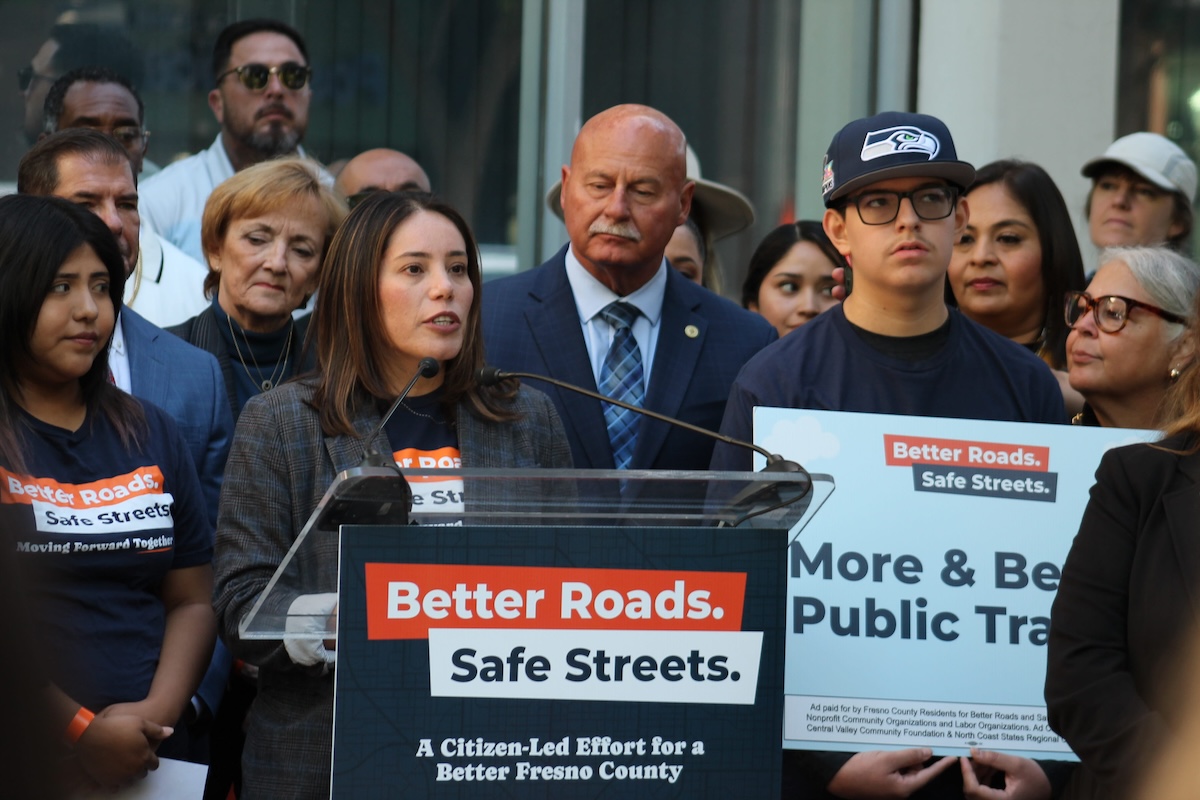 A woman wearing a plaid blazer over a navy campaign t-shirt speaking at the "Better Roads. Safe Streets." podium. She is surrounded by the same crowd of supporters and campaign members, who are watching her speak.