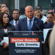 An older man with a mustache, wearing a blue suit and a patterned blue tie, speaking at the campaign podium. The woman in the plaid blazer and other supporters stand behind him listening.