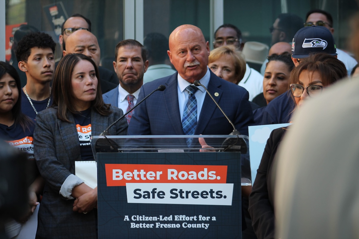 An older man with a mustache, wearing a blue suit and a patterned blue tie, speaking at the campaign podium. The woman in the plaid blazer and other supporters stand behind him listening.