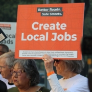 A close-up of a crowd where an older person with gray hair and sunglasses is holding up a bright orange sign. The sign reads "Better Roads. Safe Streets." at the top and "Create Local Jobs" in large white letters.