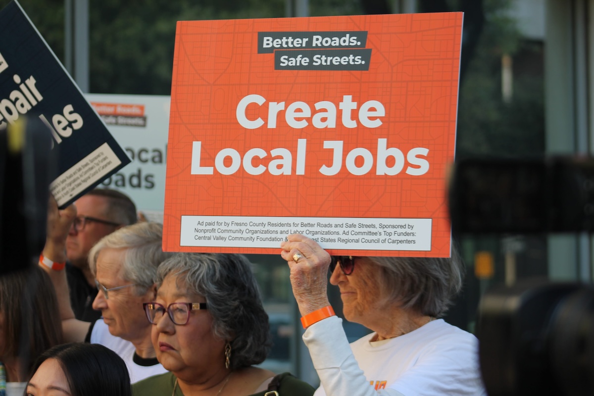 A close-up of a crowd where an older person with gray hair and sunglasses is holding up a bright orange sign. The sign reads "Better Roads. Safe Streets." at the top and "Create Local Jobs" in large white letters.