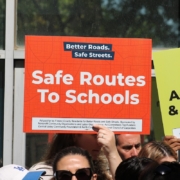 A crowd of people outdoors holding up campaign signs. Prominently featured in the center is an orange sign that reads "Safe Routes To Schools" alongside a partial view of a green sign mentioning "Strong Accountability."