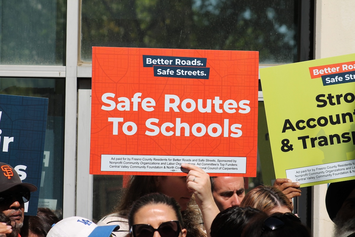 A crowd of people outdoors holding up campaign signs. Prominently featured in the center is an orange sign that reads "Safe Routes To Schools" alongside a partial view of a green sign mentioning "Strong Accountability."