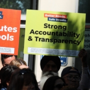 A diverse group of supporters holding up campaign signs. A bright green sign reading "Strong Accountability & Transparency" is highly visible, held next to an orange "Safe Routes To Schools" sign. A man wearing a black turban is visible in the center of the crowd.