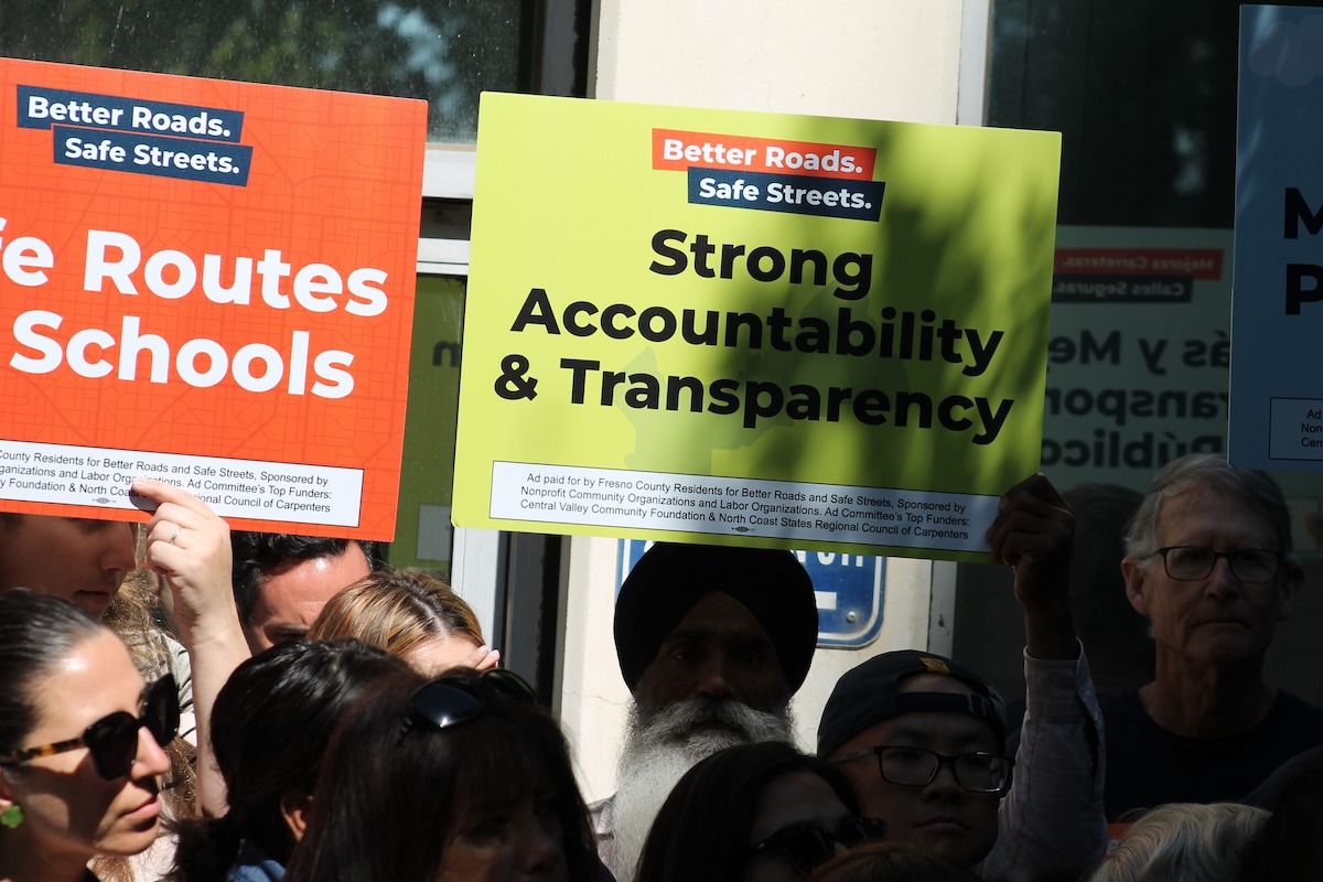 A diverse group of supporters holding up campaign signs. A bright green sign reading "Strong Accountability & Transparency" is highly visible, held next to an orange "Safe Routes To Schools" sign. A man wearing a black turban is visible in the center of the crowd.