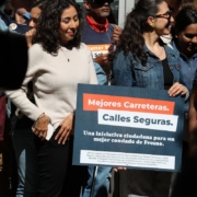 Two women standing together in a crowd. One is wearing a denim jacket and smiling while holding a dark blue campaign sign with Spanish text: "Mejores Carreteras. Calles Seguras. Una iniciativa ciudadana para un mejor condado de Fresno."