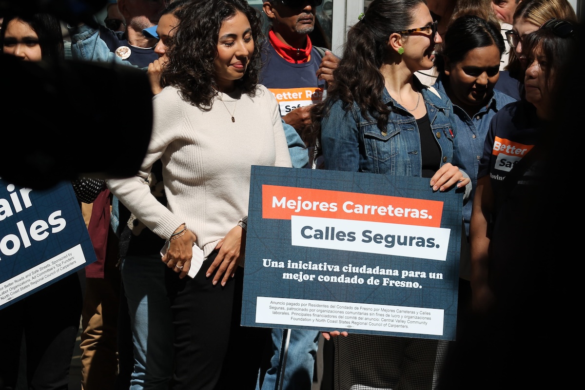 Two women standing together in a crowd. One is wearing a denim jacket and smiling while holding a dark blue campaign sign with Spanish text: "Mejores Carreteras. Calles Seguras. Una iniciativa ciudadana para un mejor condado de Fresno."