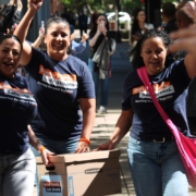 Three women wearing matching navy "Better Roads. Safe Streets." t-shirts walking through a crowd. They are carrying a cardboard Bankers Box and raising their free hands in celebration while the people around them clap and cheer.