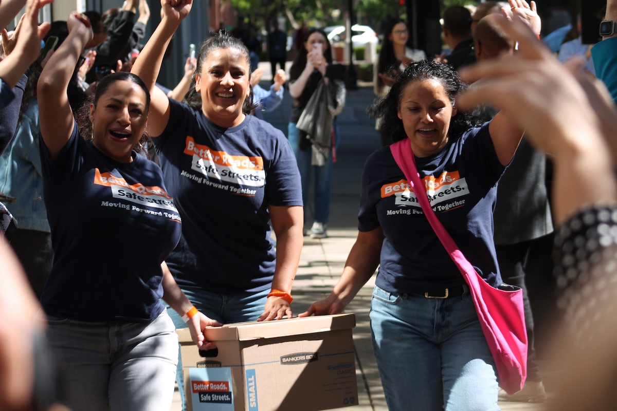 Three women wearing matching navy "Better Roads. Safe Streets." t-shirts walking through a crowd. They are carrying a cardboard Bankers Box and raising their free hands in celebration while the people around them clap and cheer.