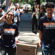 A young woman and a young man, both wearing the campaign t-shirts, are happily carrying a cardboard Bankers Box together. They are walking down a sunny sidewalk through a gauntlet of supporters who are clapping, cheering, and raising their hands to celebrate.
