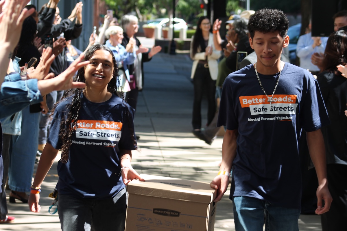 A young woman and a young man, both wearing the campaign t-shirts, are happily carrying a cardboard Bankers Box together. They are walking down a sunny sidewalk through a gauntlet of supporters who are clapping, cheering, and raising their hands to celebrate.