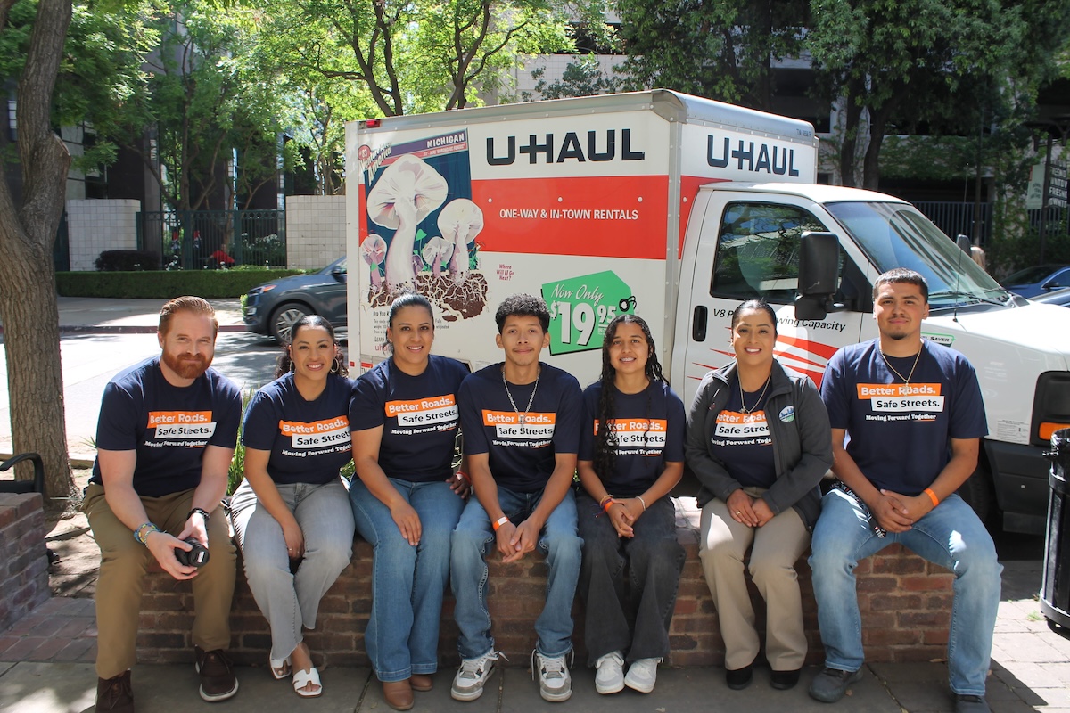 A group of seven people sitting together on a low brick wall outdoors, smiling for a group photo. They are wearing matching navy blue campaign t-shirts that read "Better Roads. Safe Streets. Moving Forward Together." Behind them is a parked white U-Haul moving truck featuring a large graphic of mushrooms on its side panel.