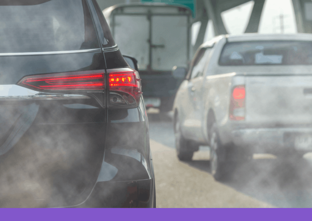 A close-up, eye-level shot from behind a black SUV in heavy traffic on a bright day. Thick, greyish-white exhaust smoke billows from the bottom of the vehicle, partially obscuring the view of a white pickup truck and a larger box truck ahead. The scene captures the visual reality of tailpipe emissions in a congested urban or highway setting, with an overhead green highway sign visible in the blurred background.
