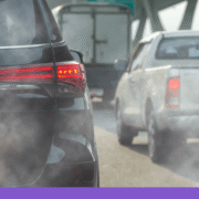 A close-up, eye-level shot from behind a black SUV in heavy traffic on a bright day. Thick, greyish-white exhaust smoke billows from the bottom of the vehicle, partially obscuring the view of a white pickup truck and a larger box truck ahead. The scene captures the visual reality of tailpipe emissions in a congested urban or highway setting, with an overhead green highway sign visible in the blurred background.