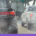 A close-up, eye-level shot from behind a black SUV in heavy traffic on a bright day. Thick, greyish-white exhaust smoke billows from the bottom of the vehicle, partially obscuring the view of a white pickup truck and a larger box truck ahead. The scene captures the visual reality of tailpipe emissions in a congested urban or highway setting, with an overhead green highway sign visible in the blurred background.