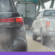 A close-up, eye-level shot from behind a black SUV in heavy traffic on a bright day. Thick, greyish-white exhaust smoke billows from the bottom of the vehicle, partially obscuring the view of a white pickup truck and a larger box truck ahead. The scene captures the visual reality of tailpipe emissions in a congested urban or highway setting, with an overhead green highway sign visible in the blurred background.