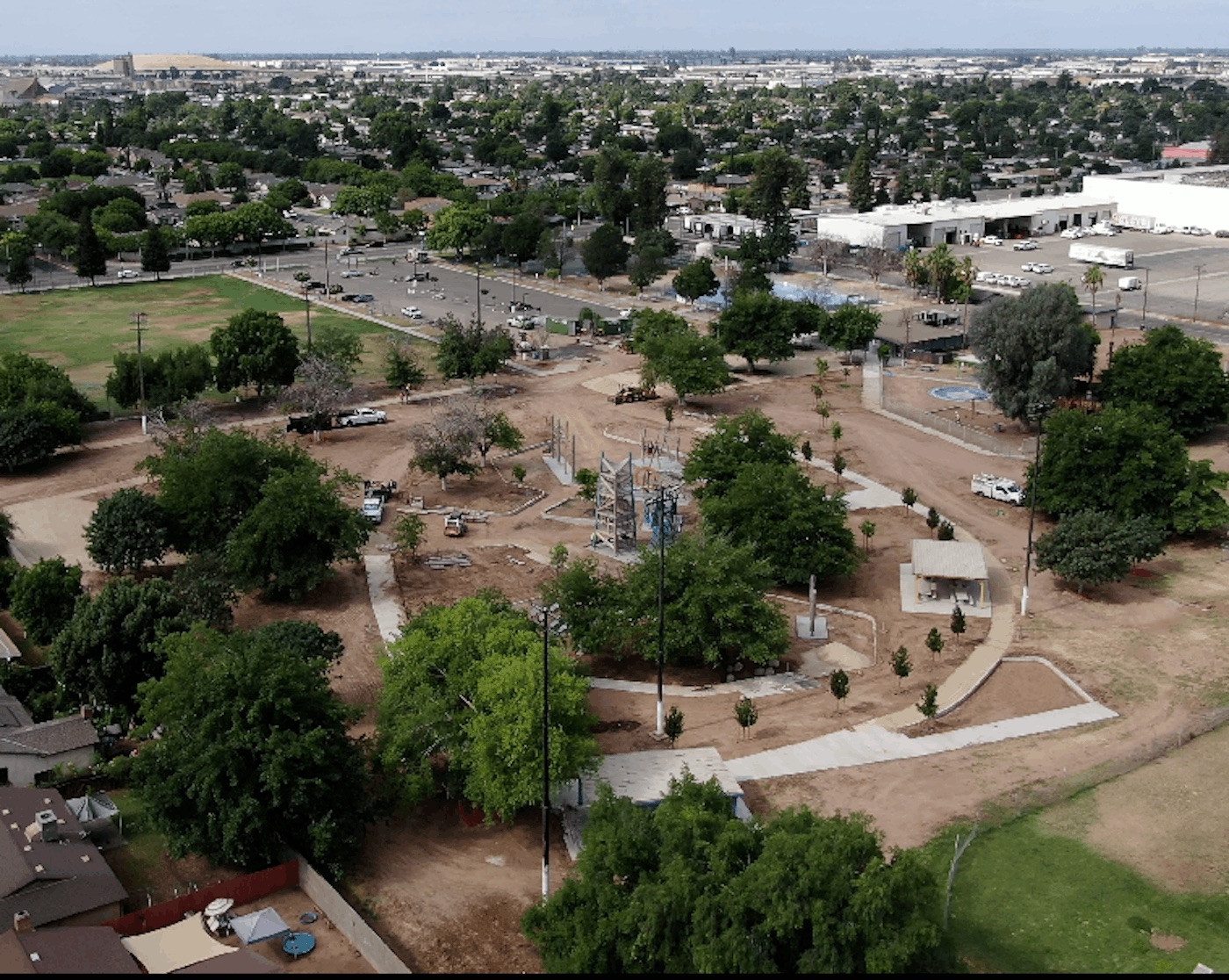 An aerial drone view looking down on the Calwa Park construction project. The site features winding concrete pathways, freshly planted trees, covered picnic structures, and diverse modern playground equipment scattered across a large expanse of graded dirt. The park is bordered by a green sports field, residential neighborhoods, and commercial buildings in the distance.