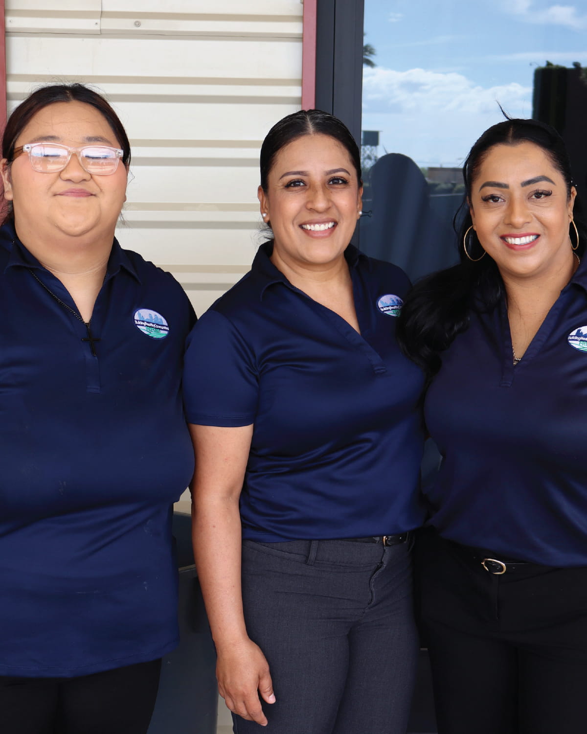 Three staff members stand side by side outdoors in front of a building, smiling at the camera. They are wearing matching navy blue polo shirts with a company logo embroidered on the chest.
