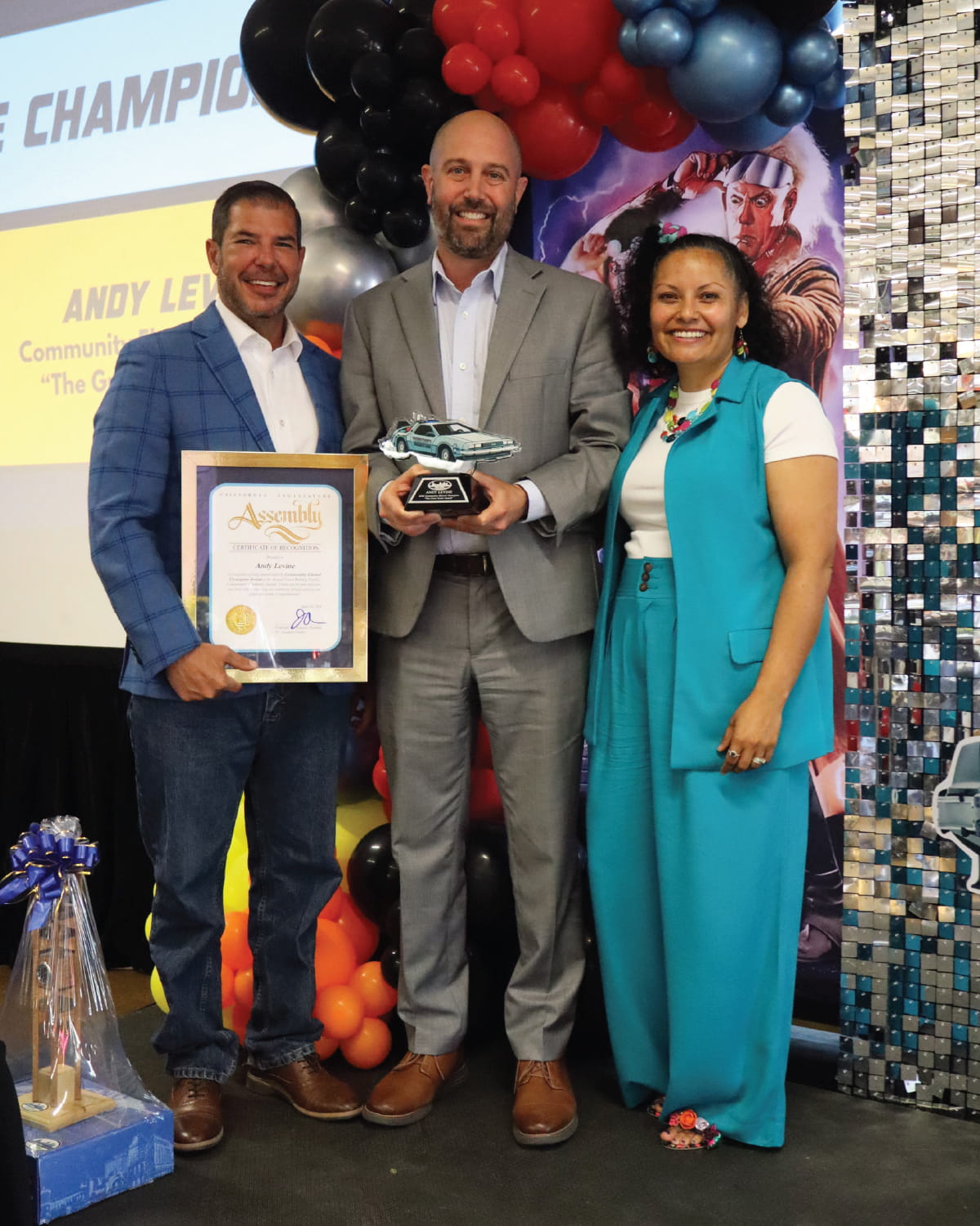 Three people pose on a stage during an awards presentation, with one person holding a certificate and another holding a trophy shaped like a car.