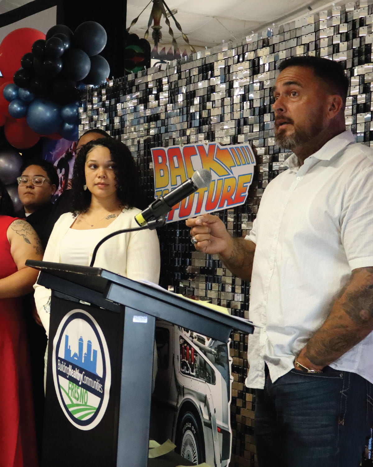 A man in a white shirt speaks at a podium with a microphone during a community event, while several attendees stand beside him. A “Back to the Future” themed backdrop and black, red, and blue balloon decorations are visible behind the group.