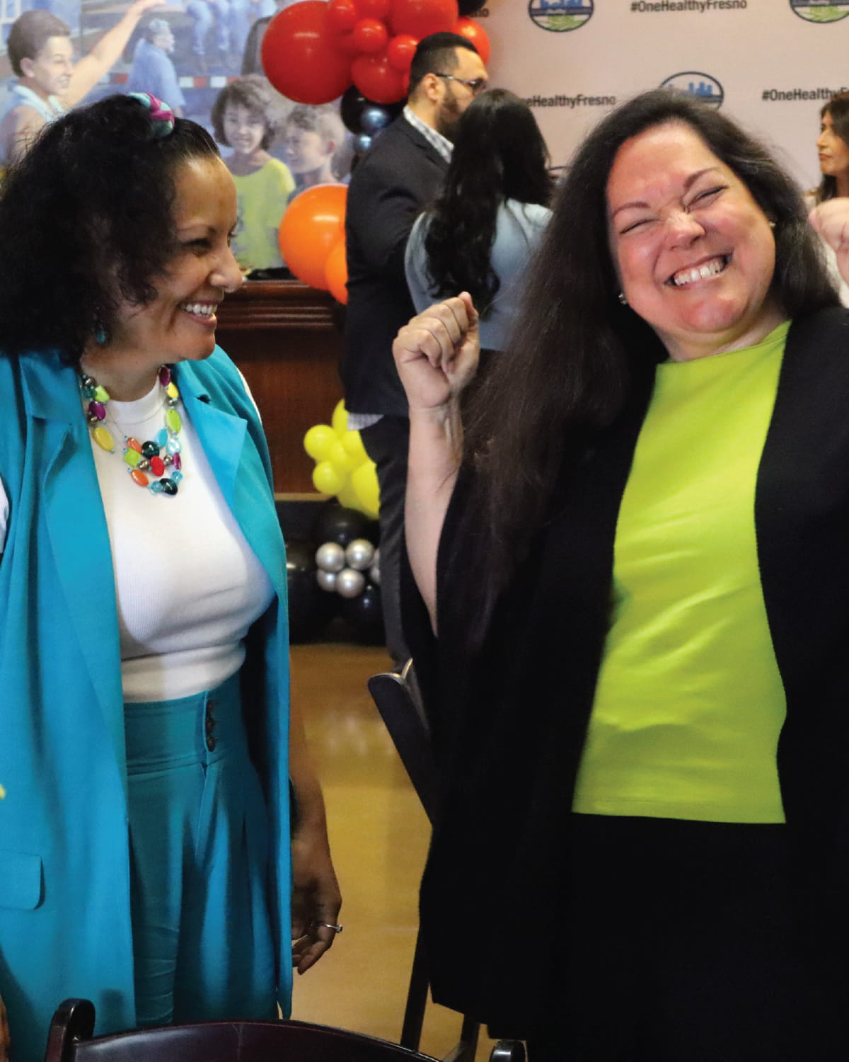 Two women share a joyous, laughing moment indoors. The woman on the right has her eyes closed in laughter and a fist excitedly raised in the air; she is wearing a bright green shirt under a black cardigan. The woman on the left smiles at her, wearing a bright teal outfit and a colorful beaded necklace. A 