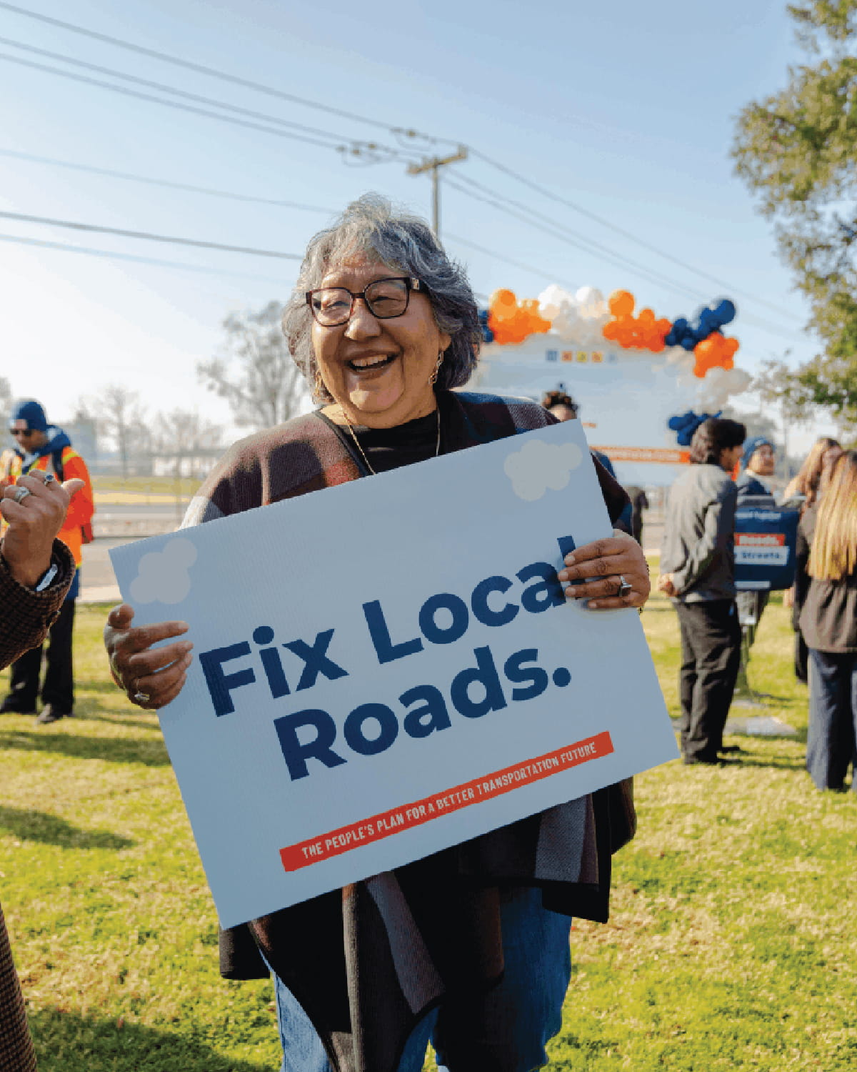 A joyful older woman with gray hair and glasses smiles brightly while holding a large sign that reads 