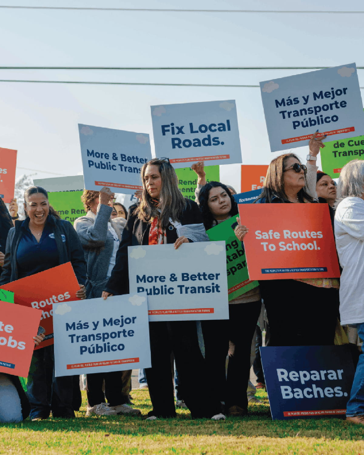 A diverse crowd of people stands on a grassy field holding up various campaign signs during a rally. The brightly colored orange, blue, and green signs feature slogans in both English and Spanish.” 
/>
<img src=