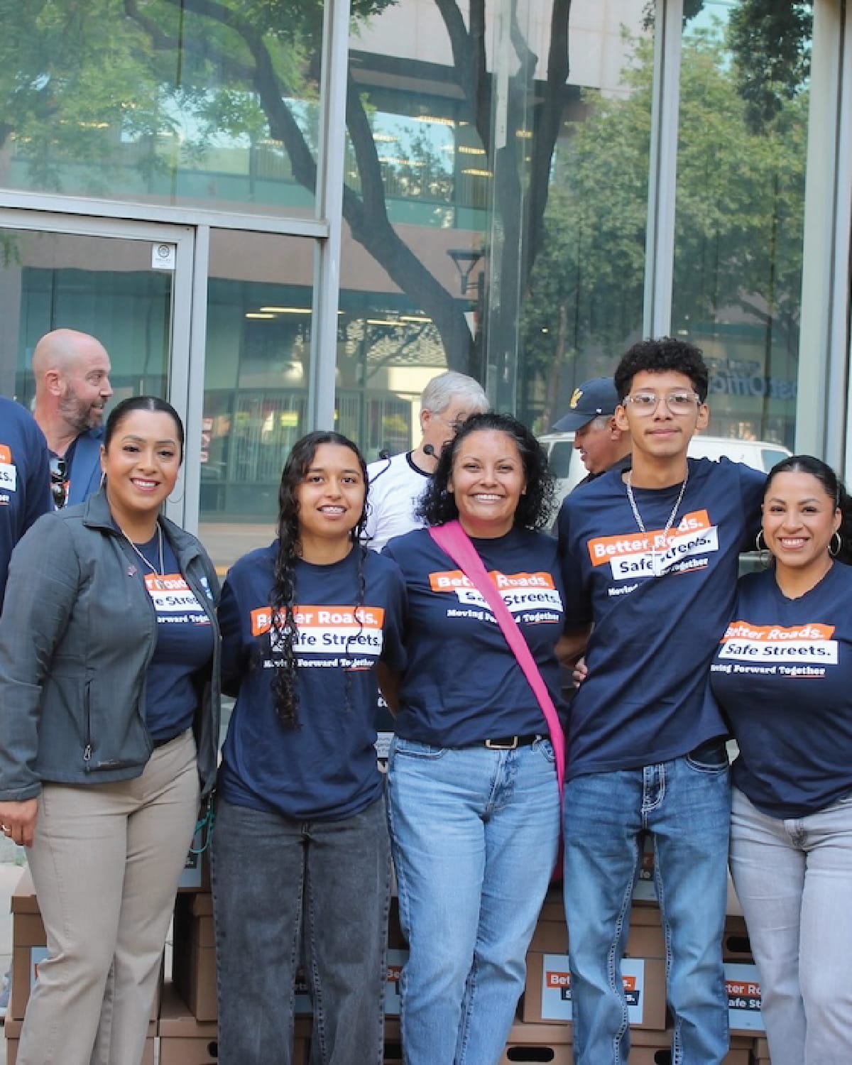 Five people stand together smiling warmly for a photo in front of a building with glass windows. Four of them are wearing matching navy blue campaign t-shirts that read 