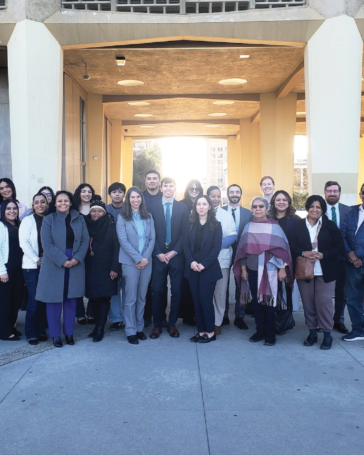 A large, diverse group of about 20 people pose for a group photograph under a concrete architectural overpass. The sun is shining brightly from behind them, casting a glow. They are dressed in a mix of professional business attire and casual cold-weather clothing.